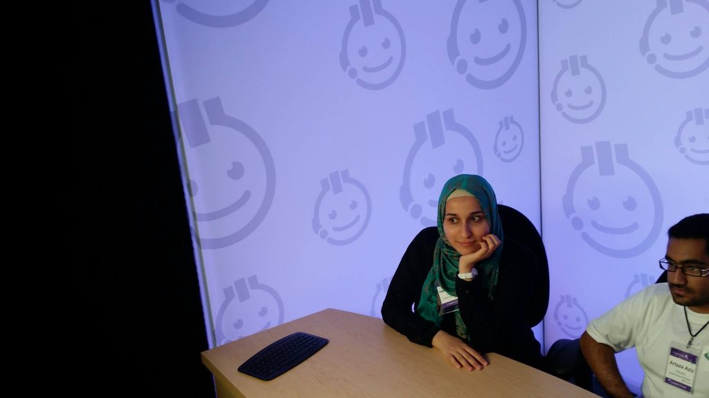 Woman in a hijab sitting at a table with a man, looking up at a screen