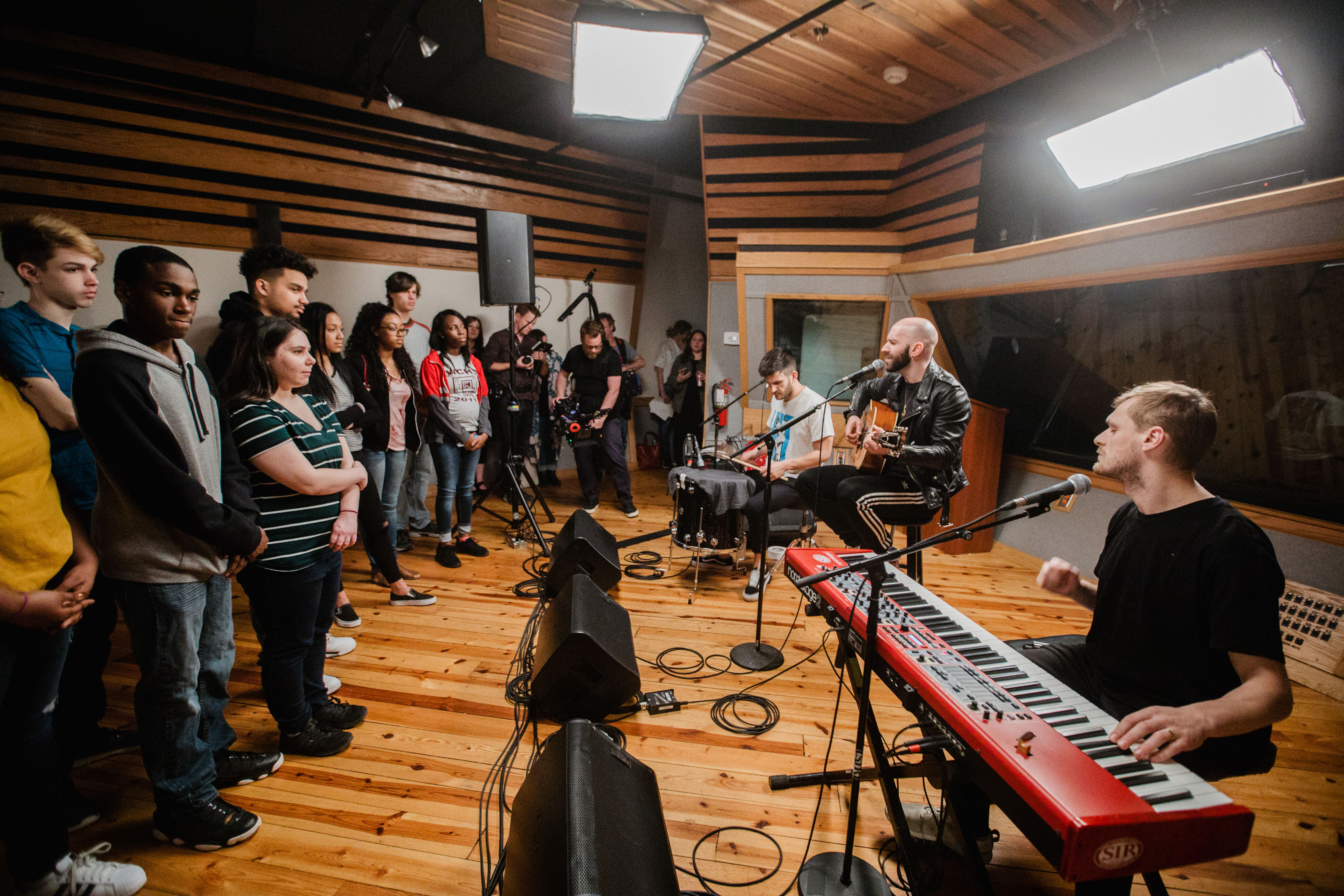 A group of students stands and watches three band members playing a song.