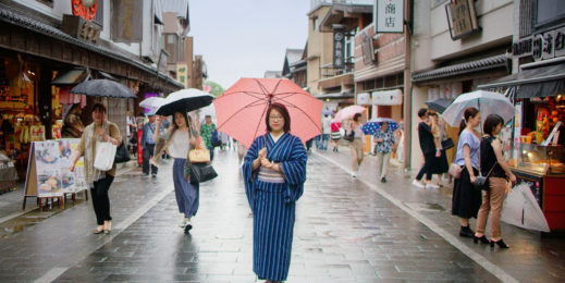 A woman wearing traditional Japanese clothing stands on a city street in Japan holding an umbrella