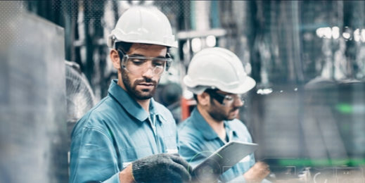 Two men in a factory wearing safety gear and interacting with mobile devices