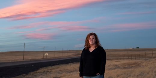 Woman stands near an open field against a blue orange sky during sunset