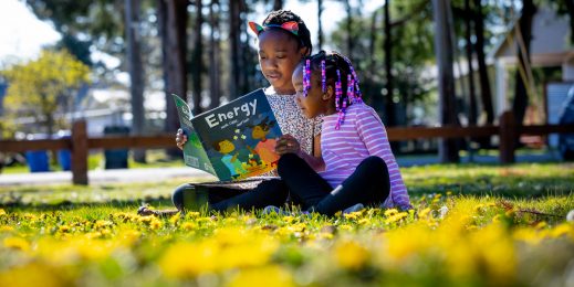 Two young girls sit close together in the grass as they read a book together