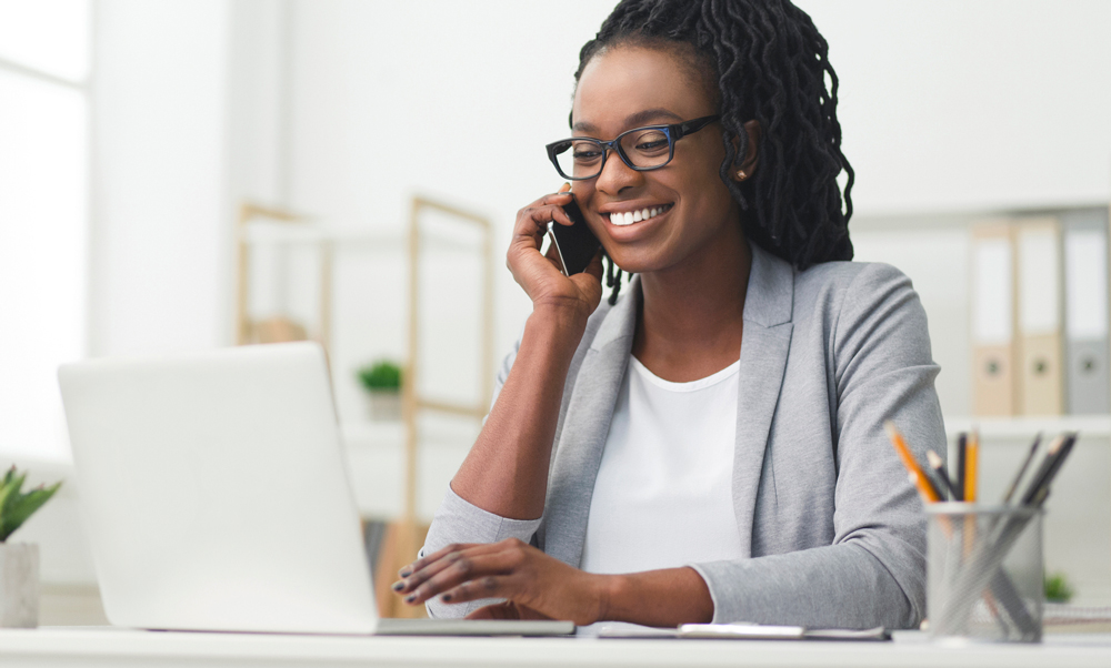 A business woman sitting at a desk, looking at a laptop and smiling while taking a phone call