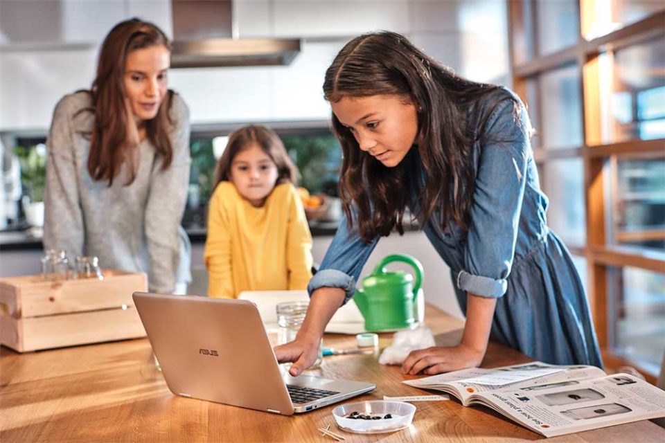 Young girl learning from PC while mom and little sister observe in their kitchen at home