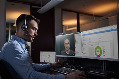 Man sitting at his desk with headphones on talking to another man on a computer monitor