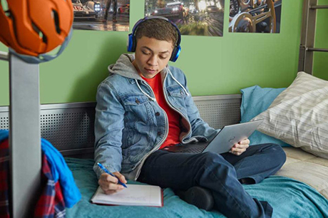 Boy sitting on bed with a laptop on his lap writing in his workbook