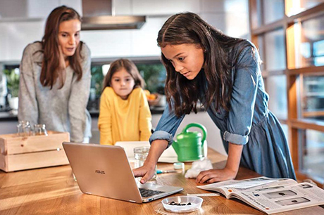 Young girl learning from PC while mom and little sister observe in their kitchen at home