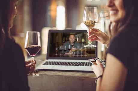 Two ladies, smiling, holding a wine glass, with a laptop open. On the Laptop screen a man is smiling while eight wine glasses are in front of him.