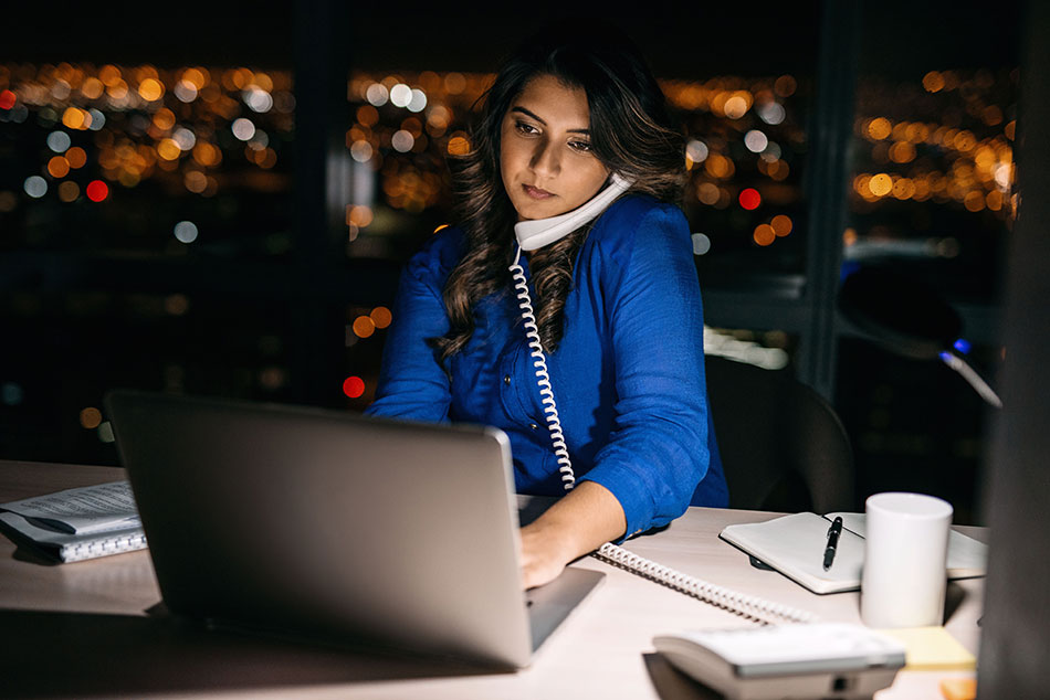 Woman working at night, using a desk phone and laptop