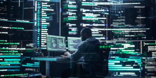 A man sits at his desk surrounded by screens that show lines of computer code.