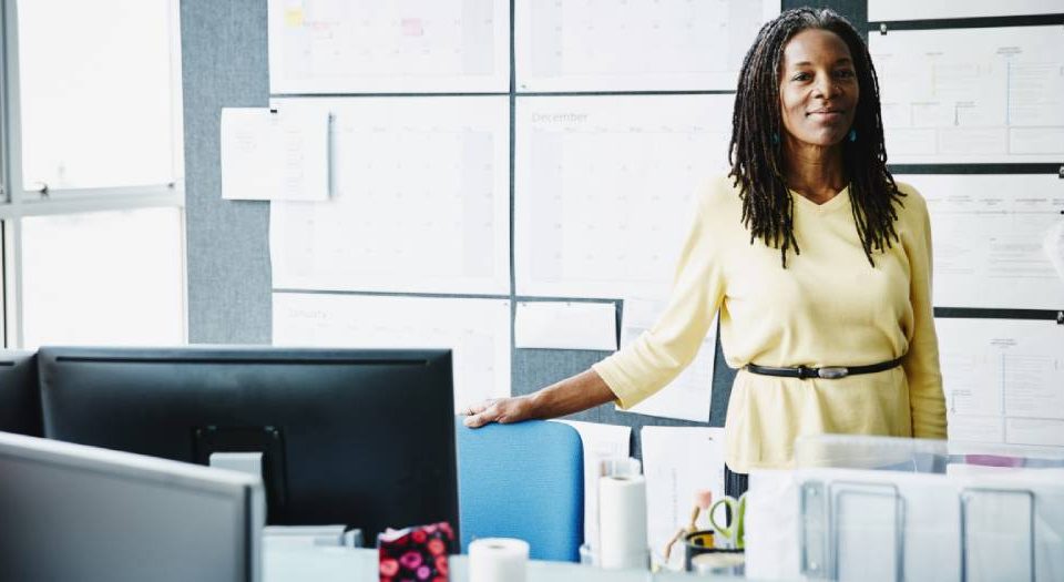Confident businesswoman standing behind an office desk, in front of wall with paper notes in the background