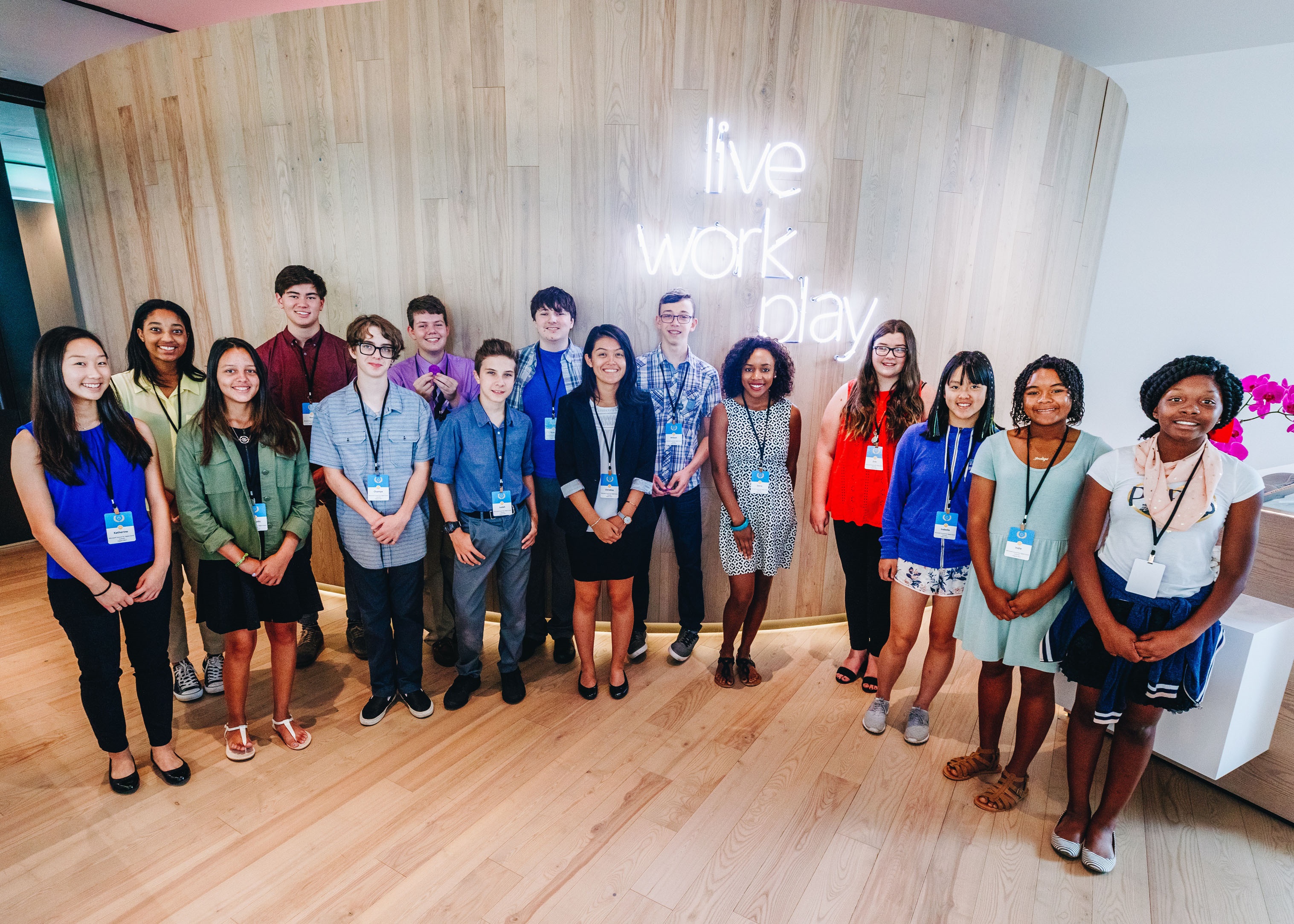 Photo of 15 teenagers standing in a semicircle in front of a neon sign saying live, work play