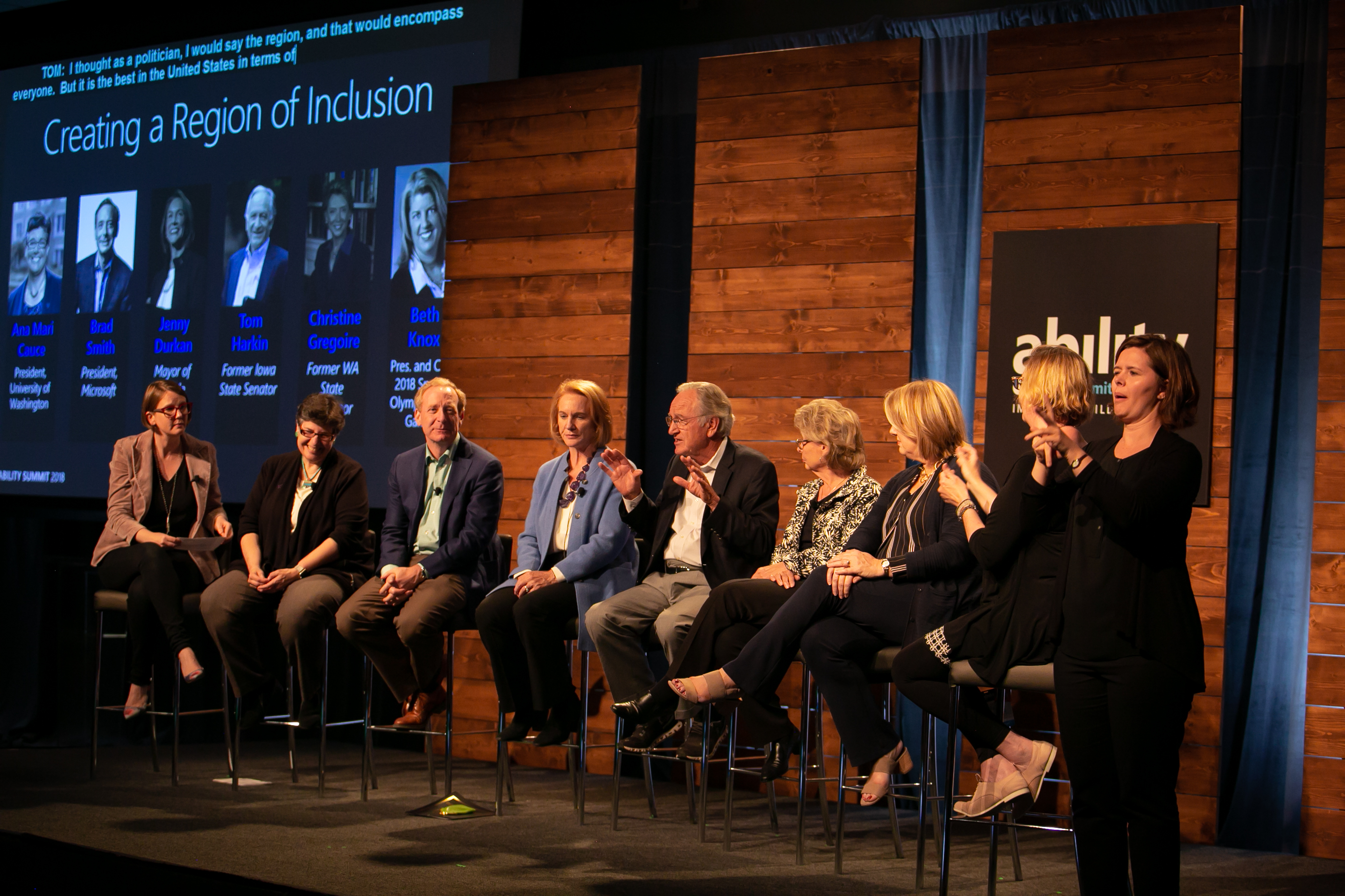 Photo of seven people sitting on chairs on stage and two interpreters signing.