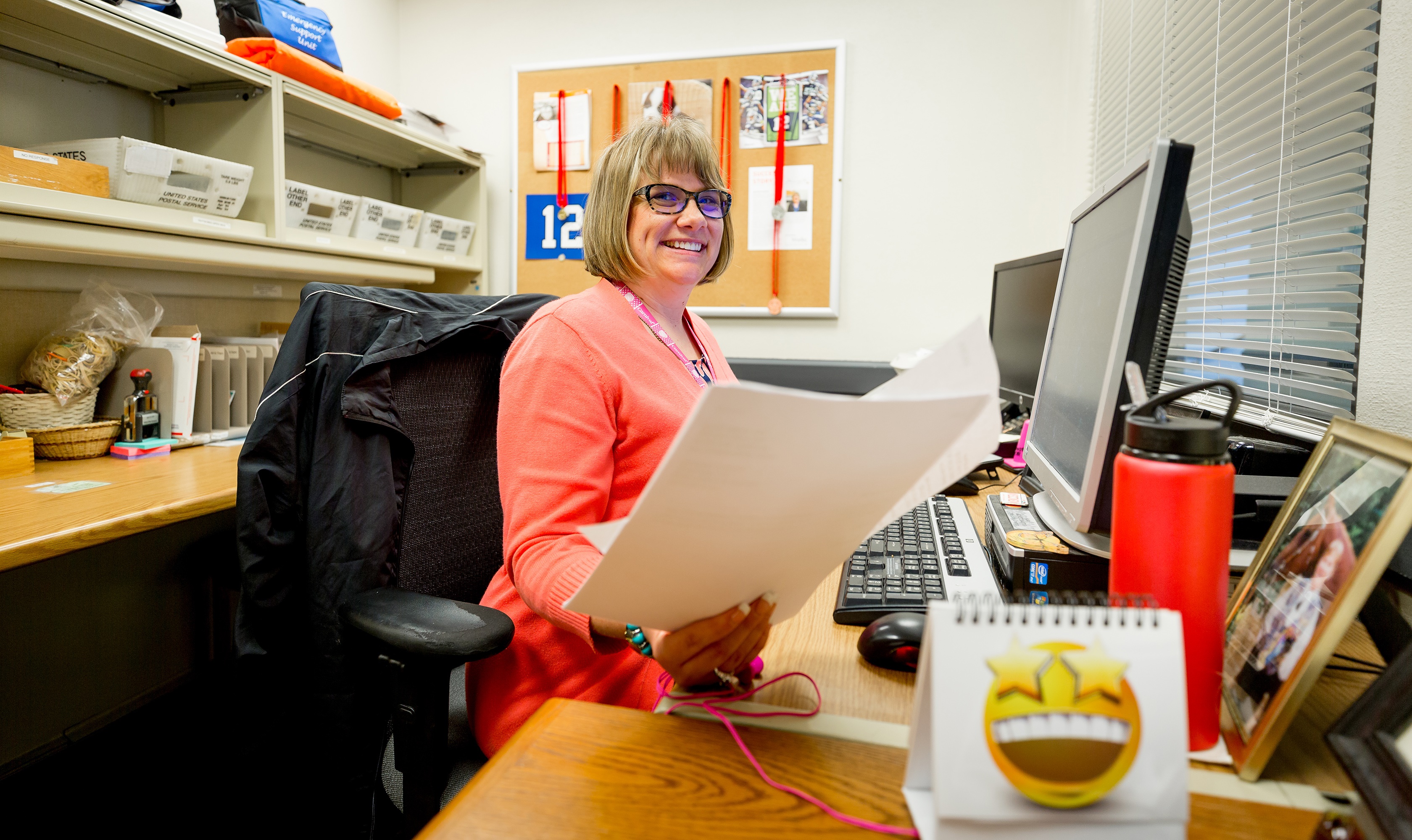 Photo of woman in front of a computer holding a piece of paper and smiling.