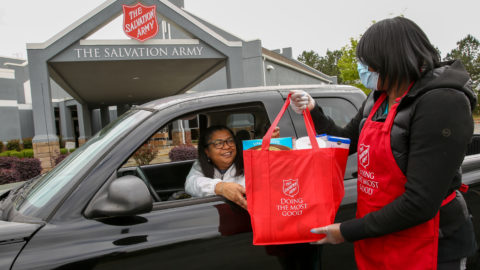 Woman in Salvation Army apron and mask handing off a bag of food to another woman in a car