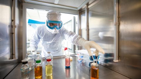 A scientist wearing a white protective suit and head covering reaches a gloved hand into an air-tight hood containing bottles of medicine.