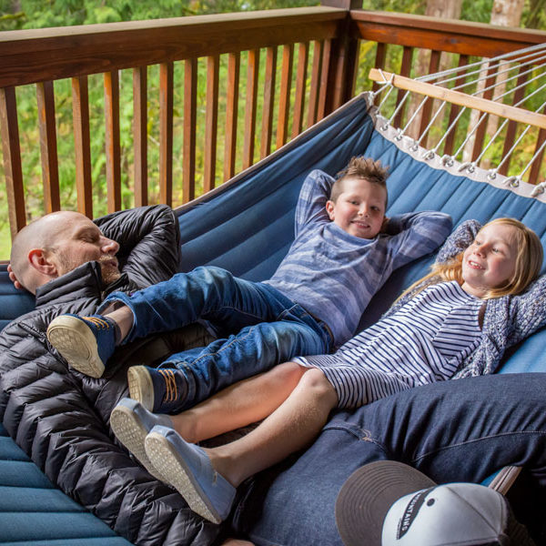 A man lays on a hammock with his children, a boy and a girl, who hang their legs over his lap