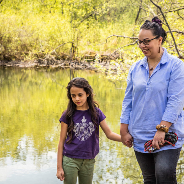 A mother and daughter hold hands by the river and smile