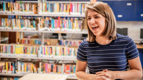 A woman smiling while standing in a school library with a shelf of books behind her