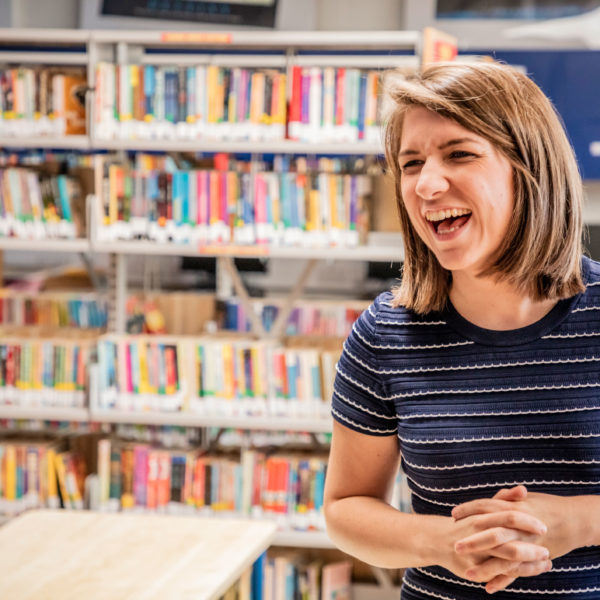 A woman smiling while standing in a school library with a shelf of books behind her