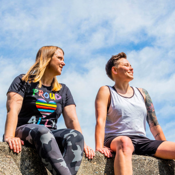 Two women sit on a stone structure at a playground. They are smiling and it's a sunny day.