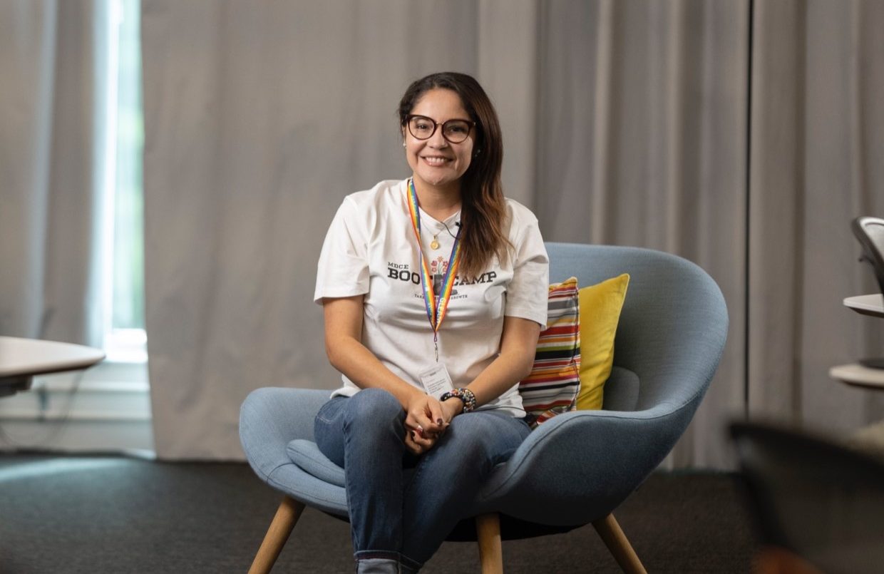 Sandra sitting in blue chair and wearing a white t-shirt and blue jeans, with a colorful lanyard.