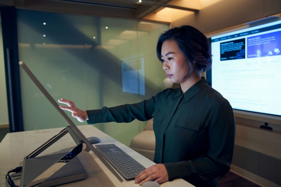 Eine Frau blickt konzentriert auf einen großen Bildschirm, den sie mit ihren Fingern berührt. Im Hintergrund ist ein großer Monitor mit Daten zu sehen.