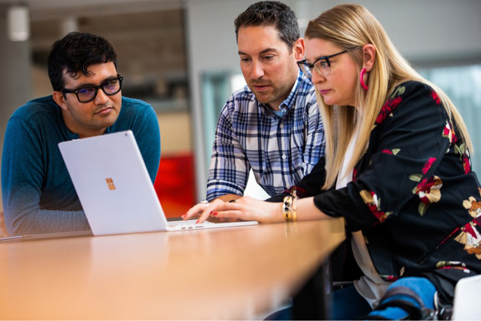 Eine Frau und zwei Männer sitzen an einem Tisch und arbeiten an einem Surface Book.