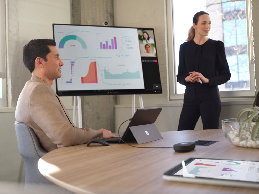 Eine Frau und ein Mann arbeiten in einem hybriden Meetingraum. Hinter ihnen steht ein Surface Hub.