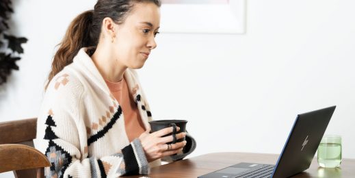 Eine Frau sitzt an einem Tisch, sie hält mit beiden Händen eine Tasse in der Hand. Vor ihr steht ein Laptop, auf den sie blickt.