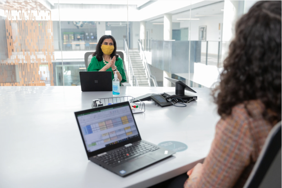 Dos mujeres con cubrebocas en una sala de juntas con laptops