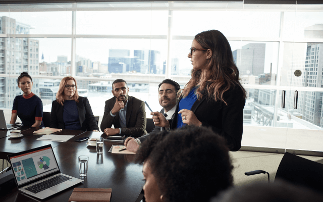 Pessoas em reunião sentadas em uma mesa e mulher apresentando