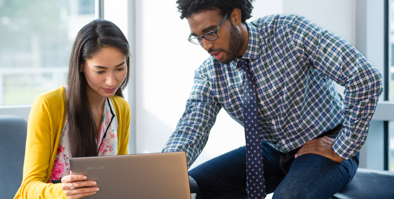 Education Color Photo of male and female staff with yellow sweater and checkered blue shirt with tie looking at device