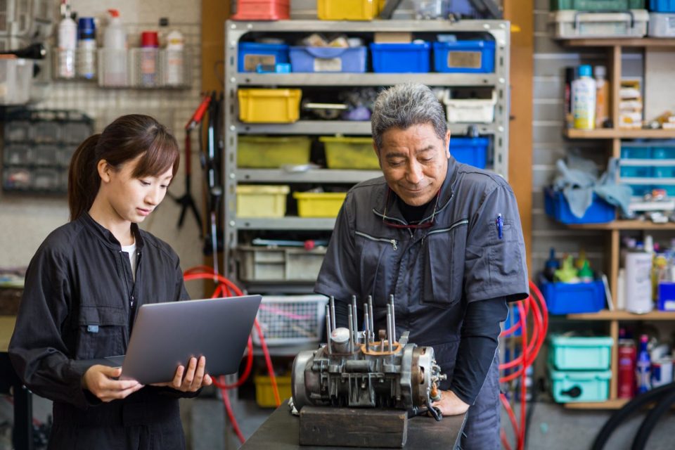 Father and daughter repairing a motorcycle engine together; daughter is using a laptop