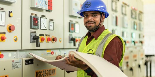 A man wearing a blue hard hat working at a machine while holding a large log book