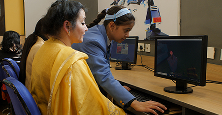 A 13 year old student in school uniform is helping her teacher with Minecraft. Her two hands are working on the keyboard while the teacher is looking at the monitor on the table. There are two other ladies and one more monitor in the picture.