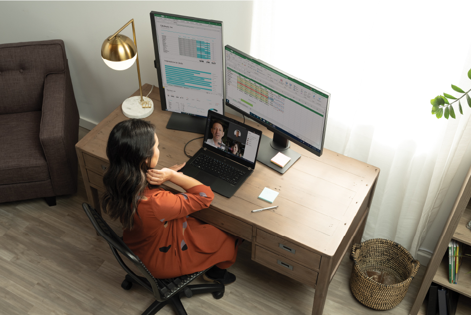 a photo of a woman with black hair and wearing an orange dress sitting in front of her laptop connected to two screens