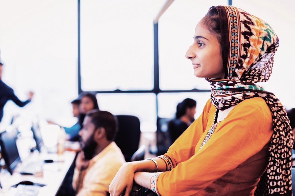 a side profile photo of a woman wearing an orange dress and scarf with people working in an office in the background