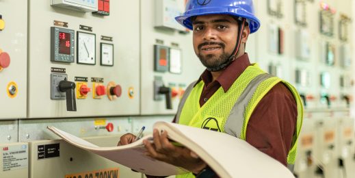 A man wearing a blue hard hat working at a machine while holding a large log book.