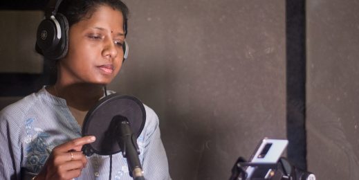 A woman reading aloud text from a smartphone’s display in front of a microphone at a recording studio.