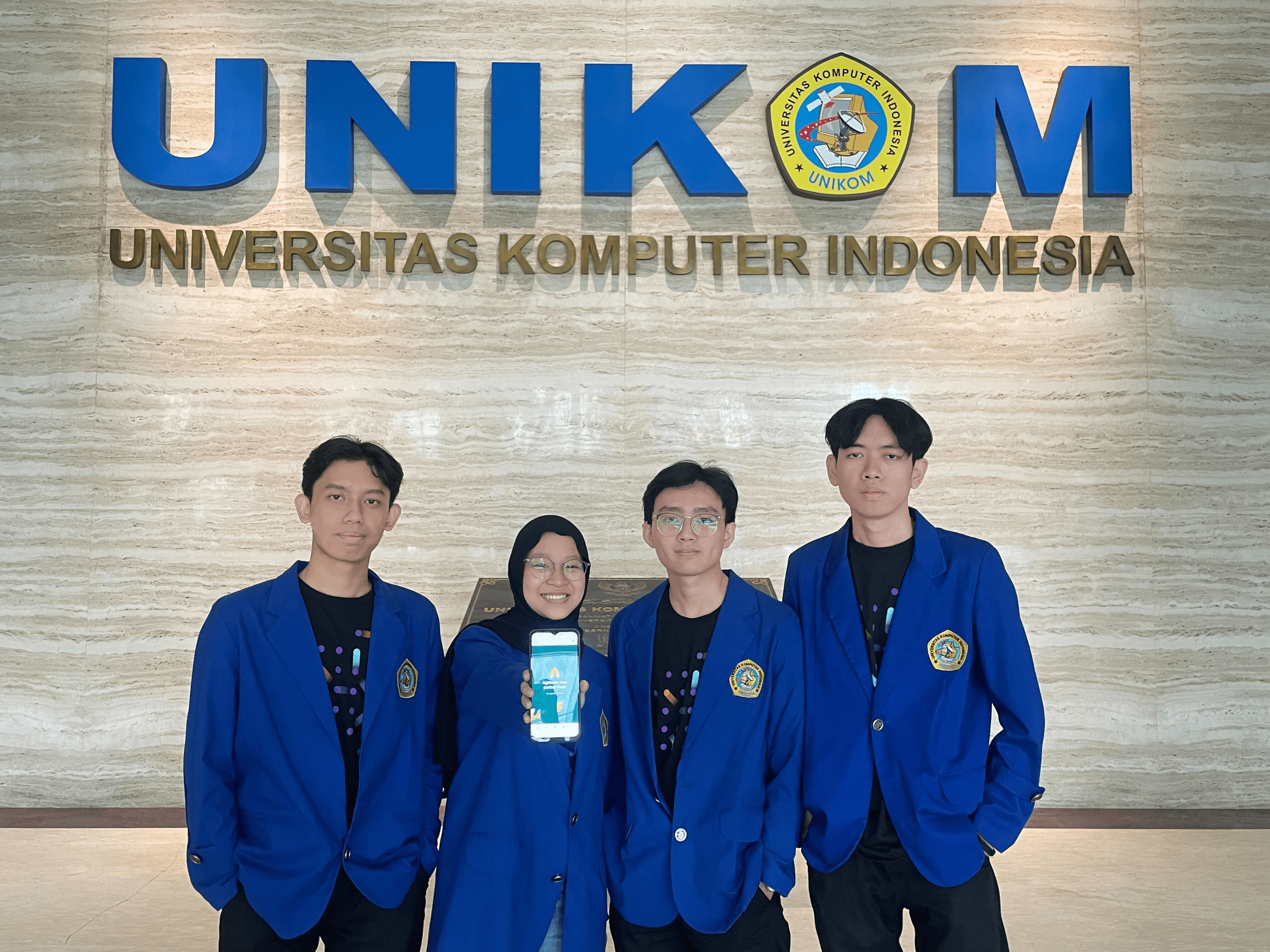 Four students in blue blazers, one of them showing a mobile app interface, standing in front of the university's logo on a wall.