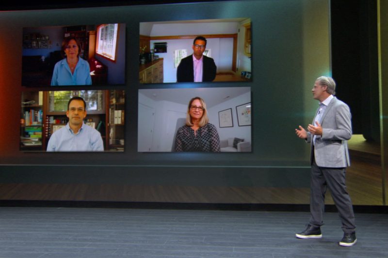 A man stands on stage speaking to a four people on a video call