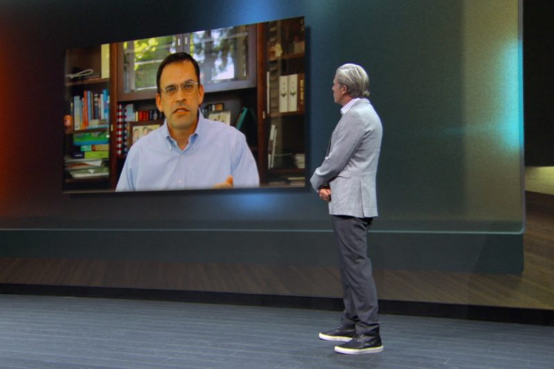 A man speaks on stage with another person on a video call