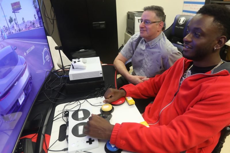 Two men play an Xbox game, one of them using an adaptive controller