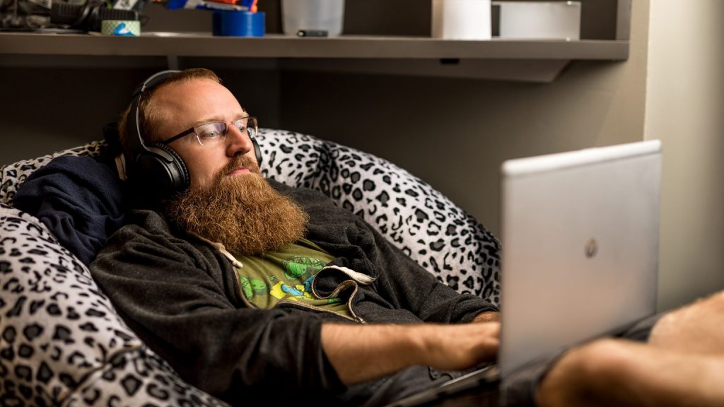 A man sits in a chair working on a laptop, with headphones on