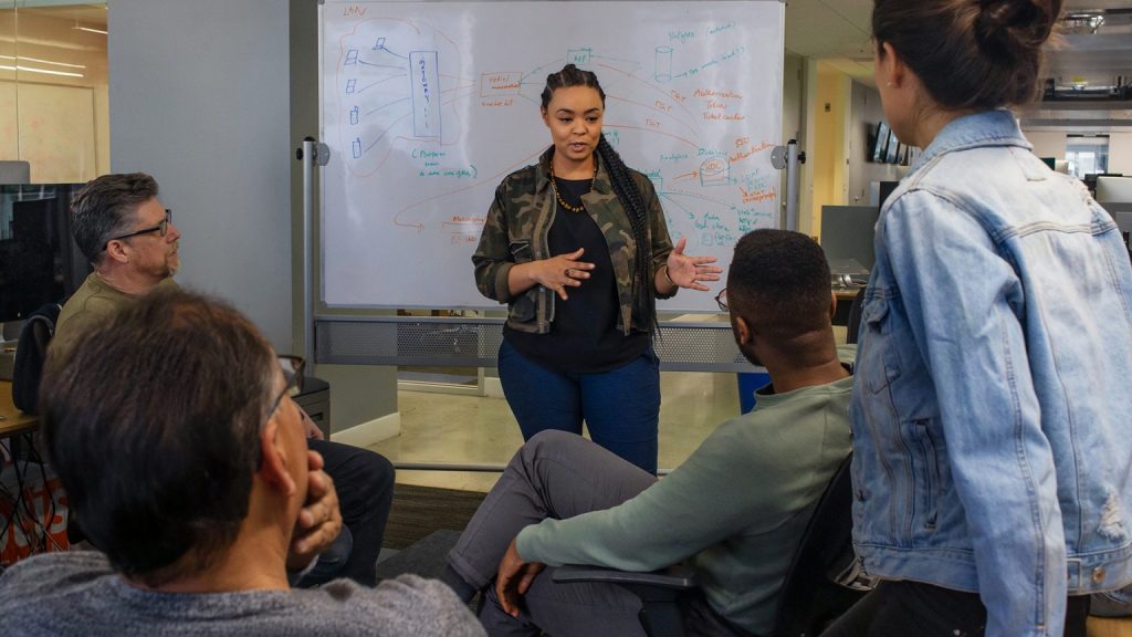 A woman leads a group of men with a whiteboard behind her, in front of men and women