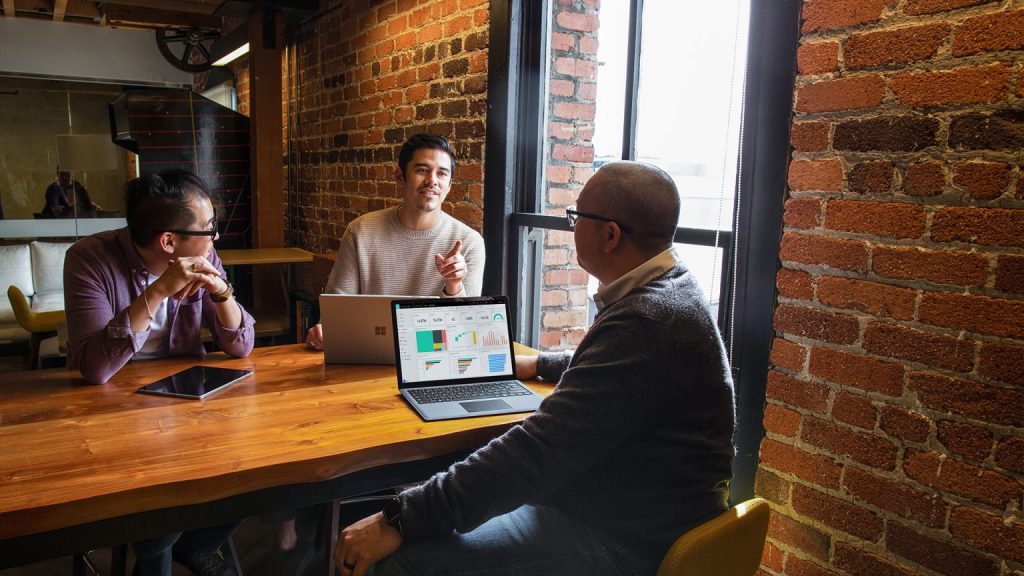 Three men sit at a desk with tablets and laptops talking