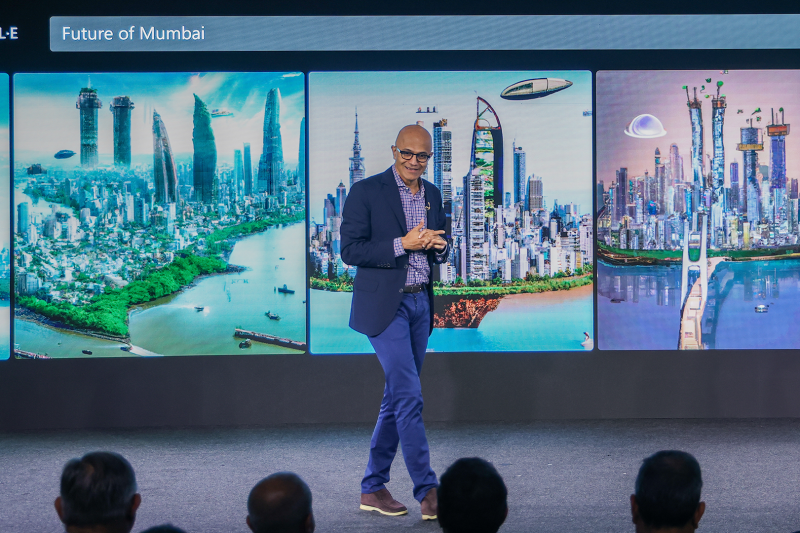 a man wearing a blue jacket delivering a keynote with the screen showing Dall-E's representation of future of Mumbai