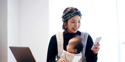 Japanese woman in casual clothes carrying her baby in baby carrier and using a smartphone on the desk.