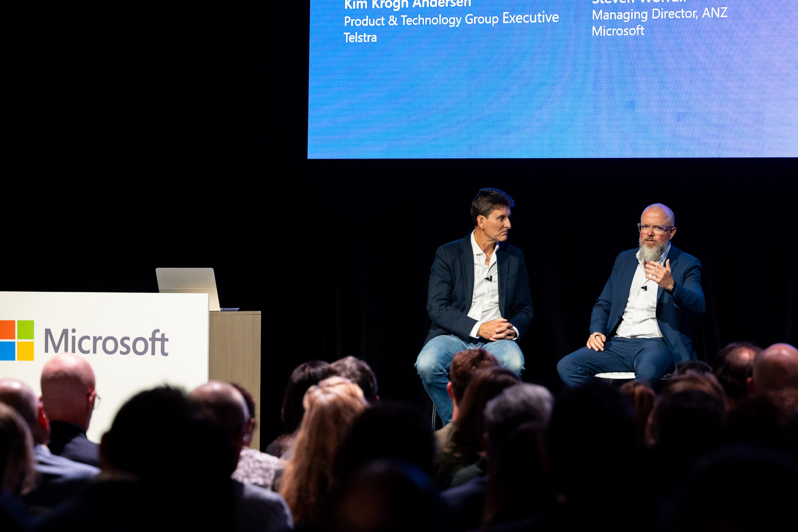 Two Caucasian men sitting on stage infront of a group of people. 